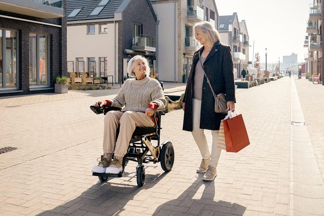 Frau in erivo Elektrorollstuhl mit Begleitung unterwegs auf Promenade.