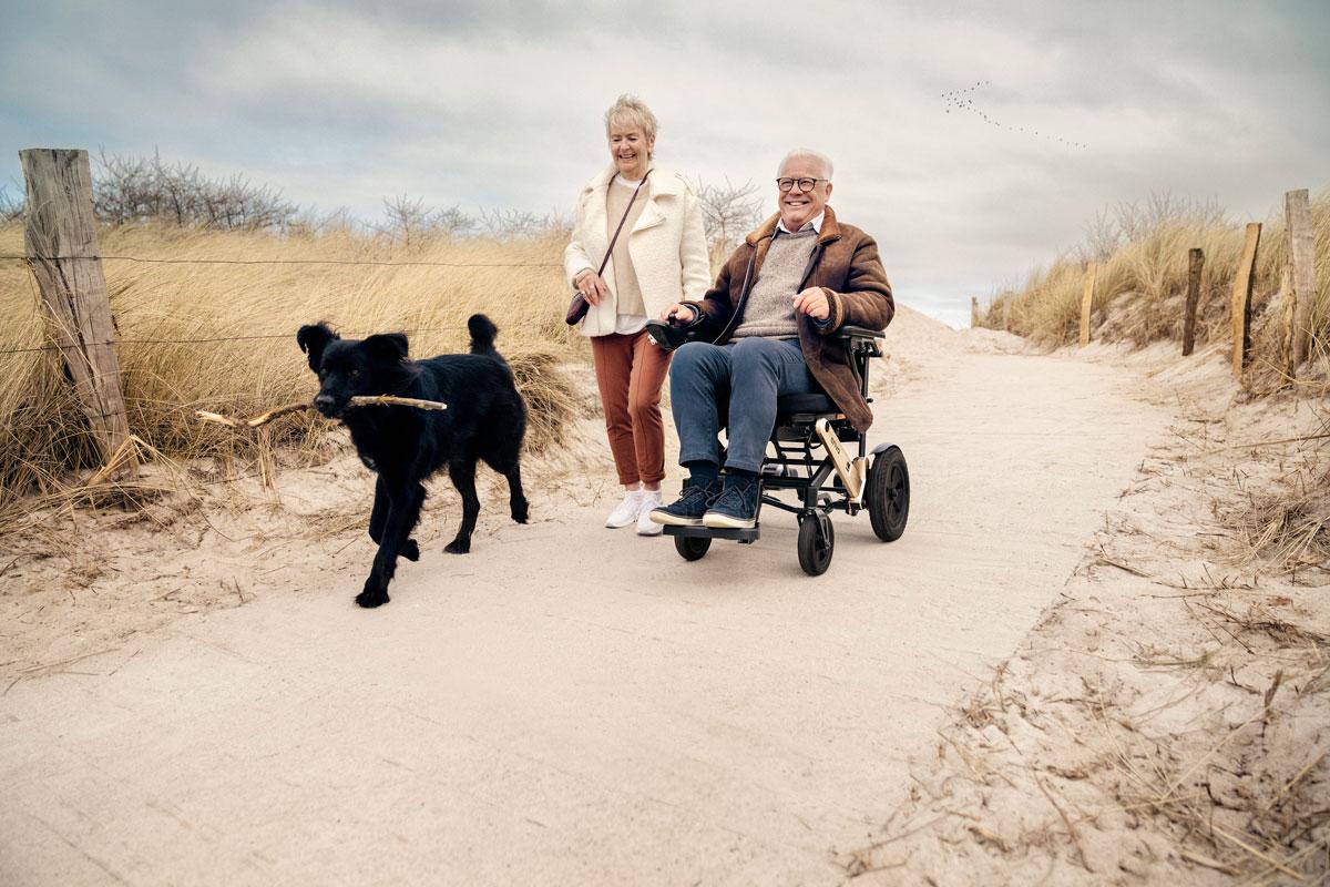 Mann in erivo Elektrorollstuhl unterwegs auf Strandweg in Begleitung von Frau und Hund.