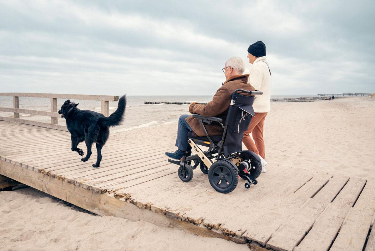 Mann in erivo Elektrorollstuhl am Strand in Begleitung von Frau und Hund.
