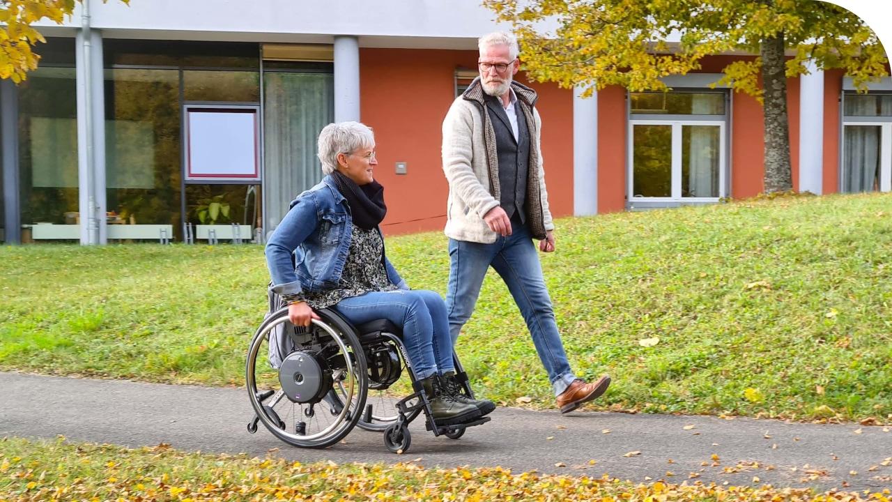 Frau fährt draußen in Elektrorollstuhl mit ihrem Mann durch die Herbstlandschaft