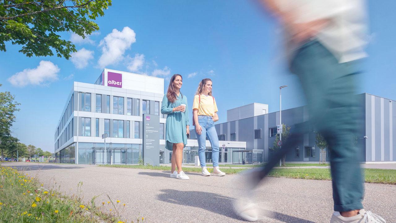Zwei Mitarbeiterinnen der Alber GmbH gehen vor dem neuen Firmengebäude bei frühlingshaftem Wetter entlang