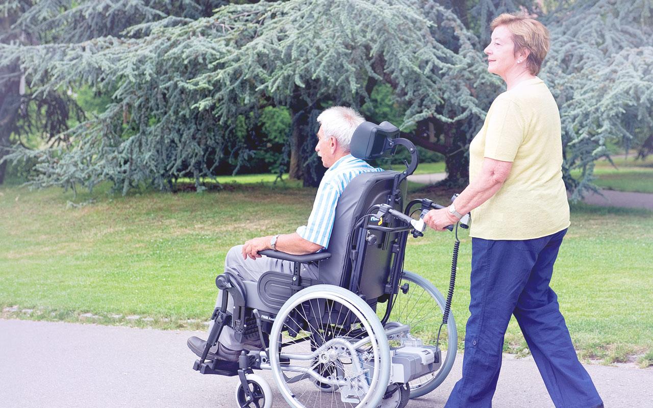 Seitlicher Blick auf Mann in Pflegerollstuhl mit viamobil Brems- und Schiebehilfe, Frau als Begleitung dahinter.