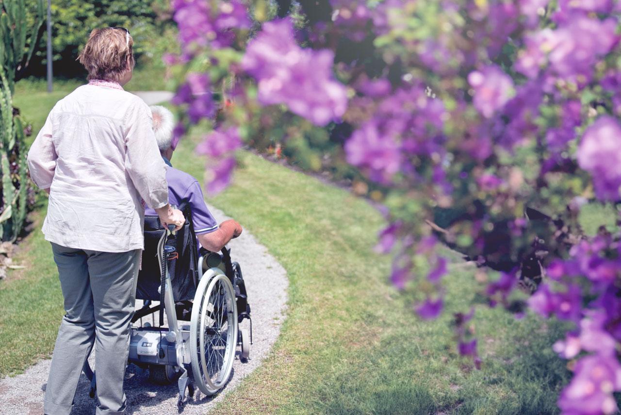 Blick durch sommerliche Blüten, Frau schiebt Mann in Rollstuhl mit viamobil Brems- und Schiebehilfe durch Park.