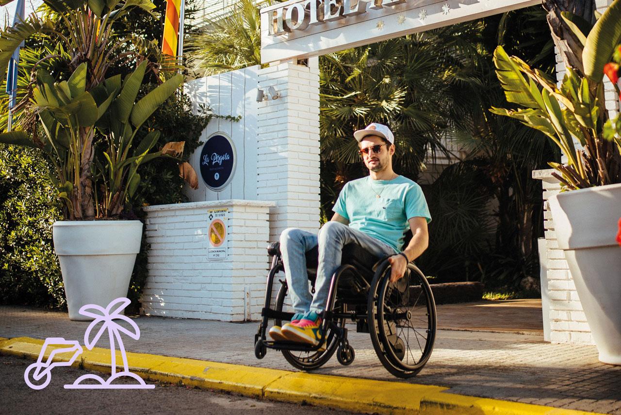 Junger Mann in Aktivrollstuhl mit smoov Elektroantrieb am Urlaubsort mit vielen Grünpflanzen vor Hotel.