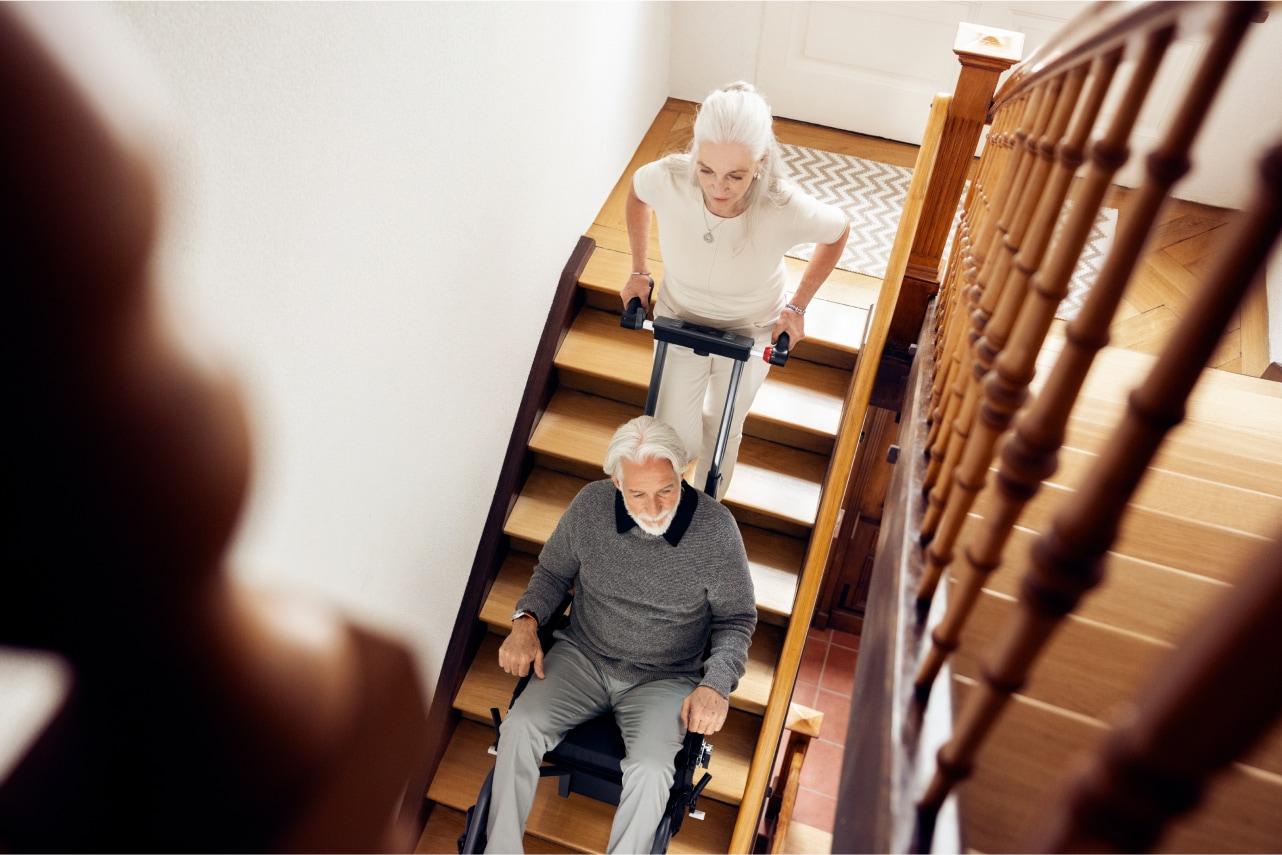 Frau begleitet Mann mit mobilem Treppensteiger scalamobil die Treppen im Haus hinunter.