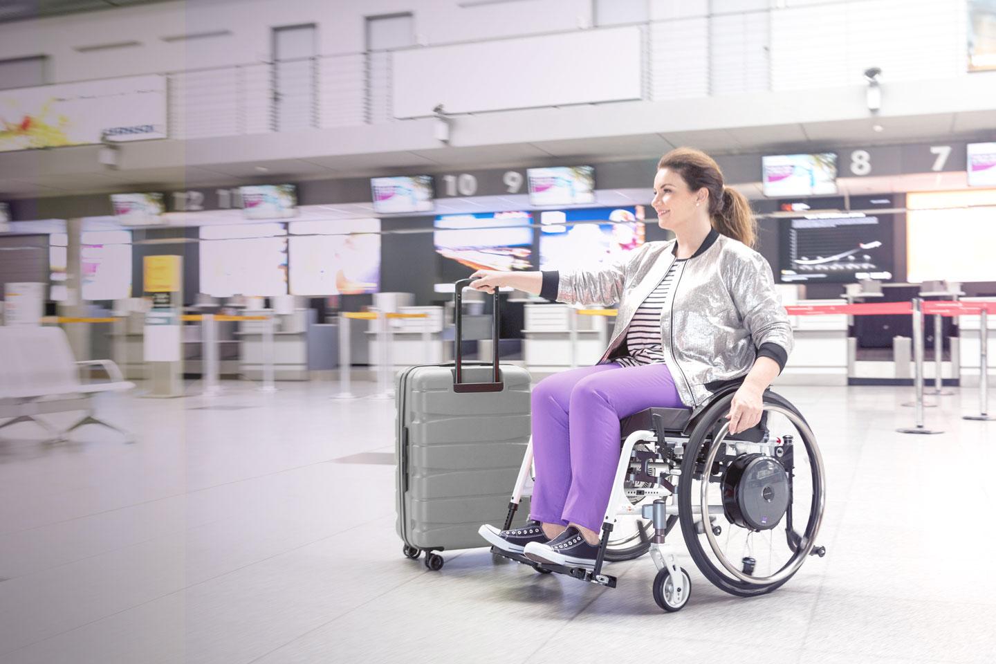 Frau im Rollstuhl mit e-motion Zusatzantrieb im Bereich der Check-In Schalter am Flughafen mit Koffer