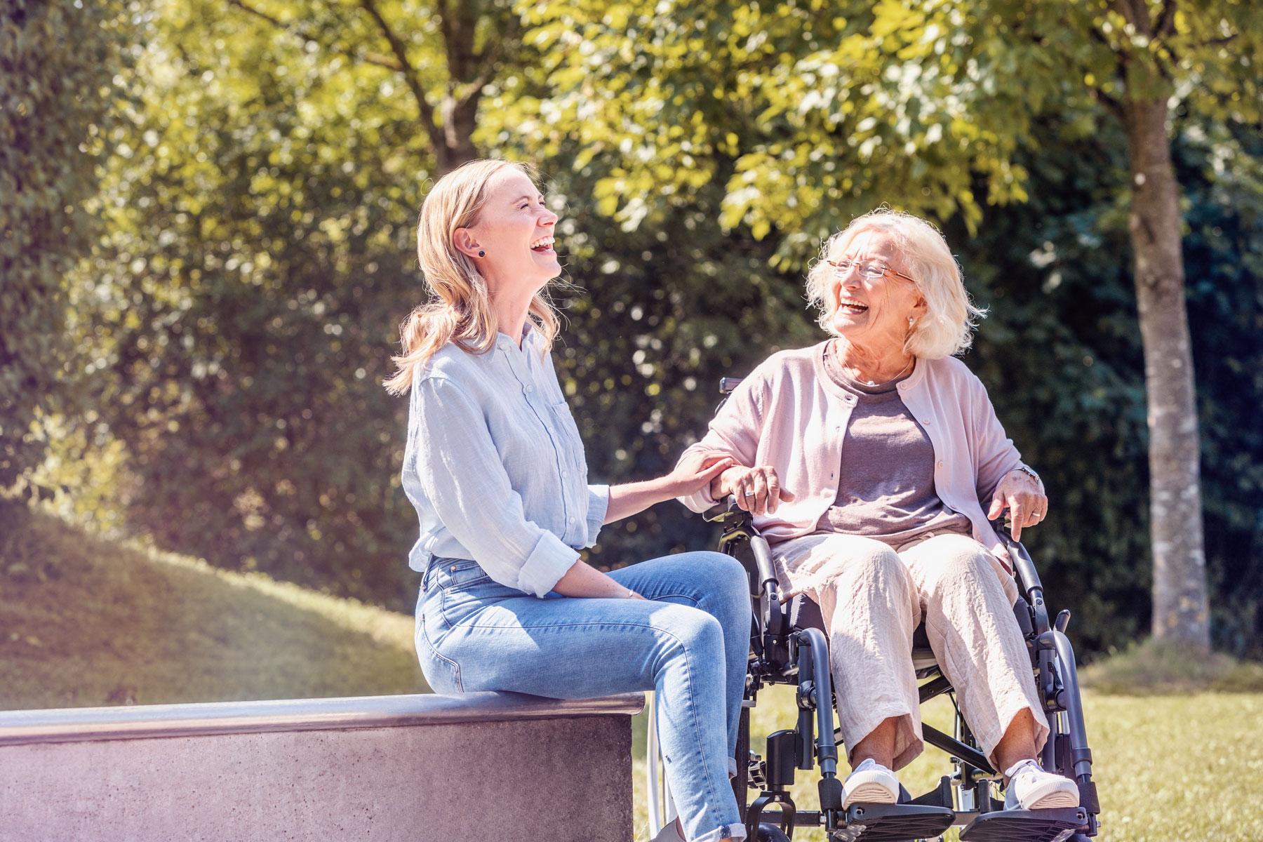 Frau in Rollstuhl mit Brems- und Schiebehilfe unterhält sich lachend mit Tochter während einer Pause im Park