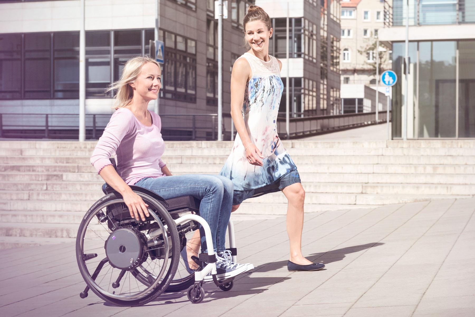 Junge Frau begleitet Freundin im Rollstuhl mit kraftverstärkendem Antrieb e-motion durch die Stadt.