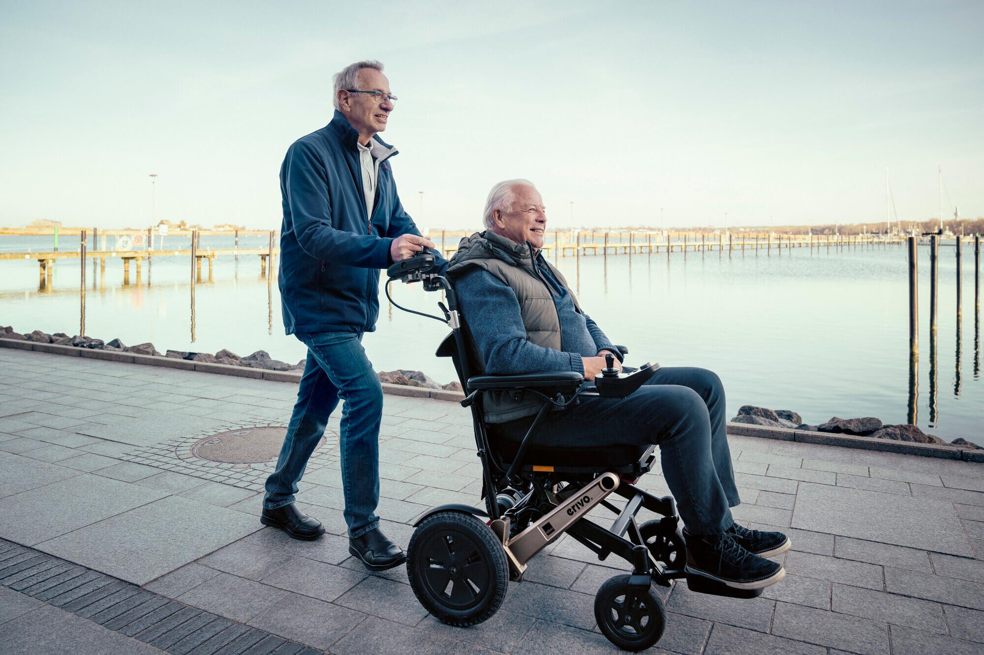 Mann fährt mit Elektrorollstuhl in Begleitung eines Freundes lächelnd am Hafen entlang