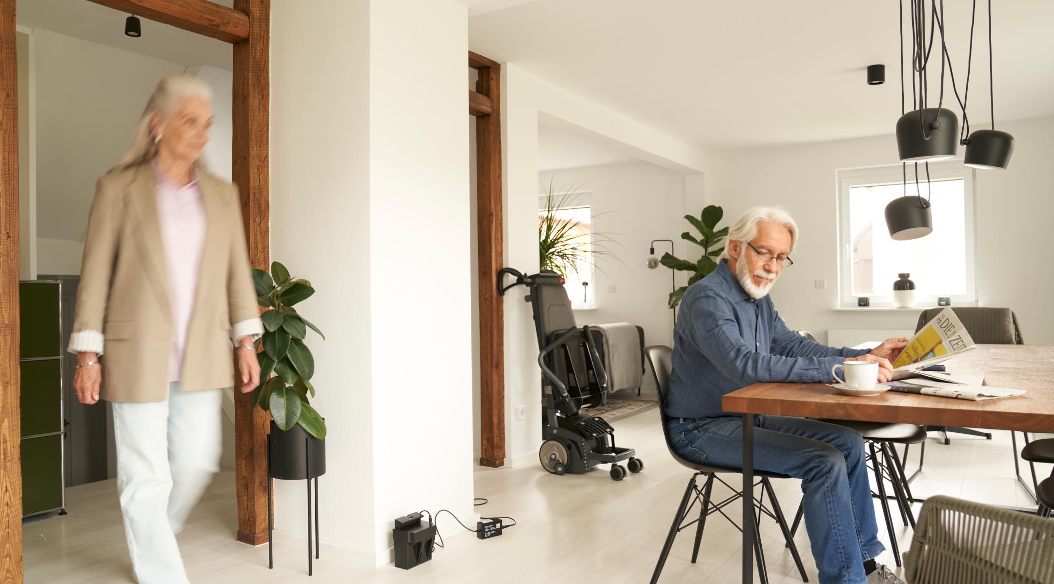 scalacombi steht zusammengeklappt an der Wand während Mann am Tisch sitzend liest und Frau die Treppen herunter kommt