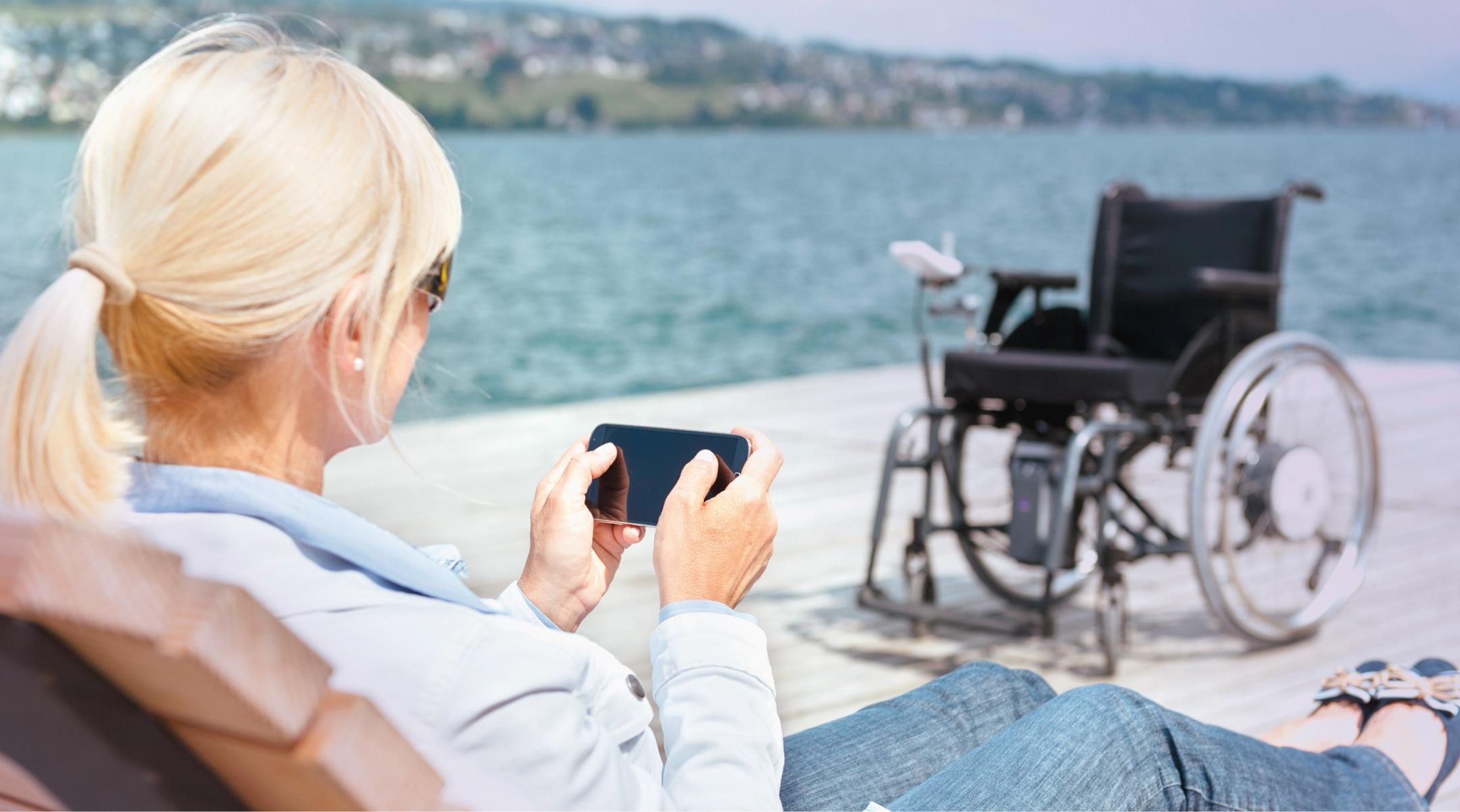 Frau im Liegestuhl mit Blick auf Smartphone, ihren Rollstuhl mit e-fix Elektroantrieb und den dahinter liegenden See