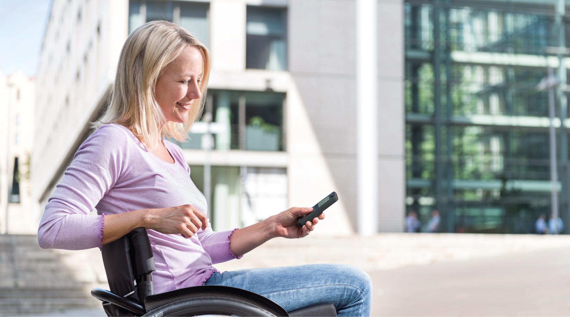 Frau in Rollstuhl mit e-motion Zusatzantrieb vor Gebäude hält ECS-Fernbedienung in der Hand