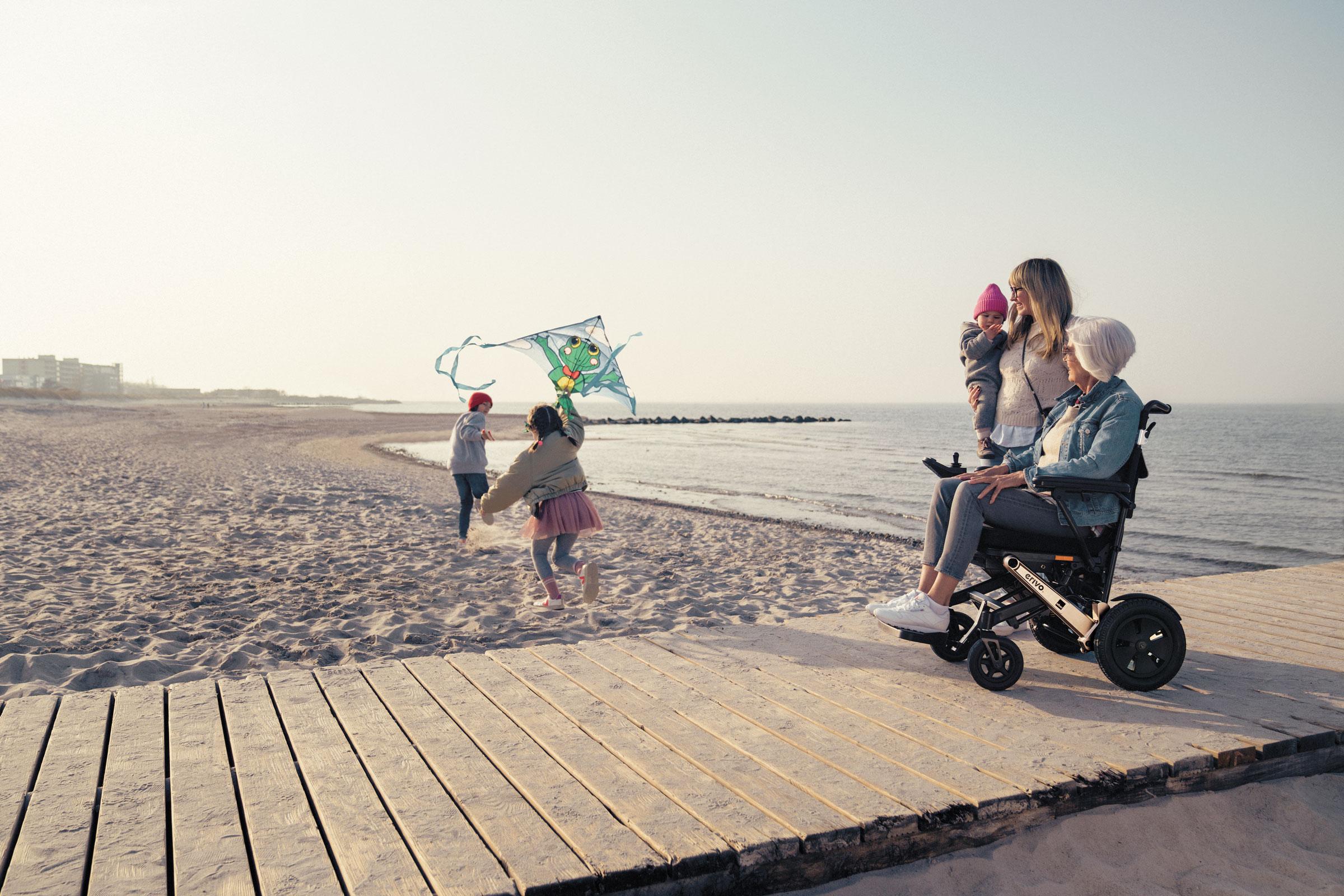 Familie und Frau in erivo Elektrorollstuhl auf Steg am Strand mit Kindern die einen Drachen steigen lassen.