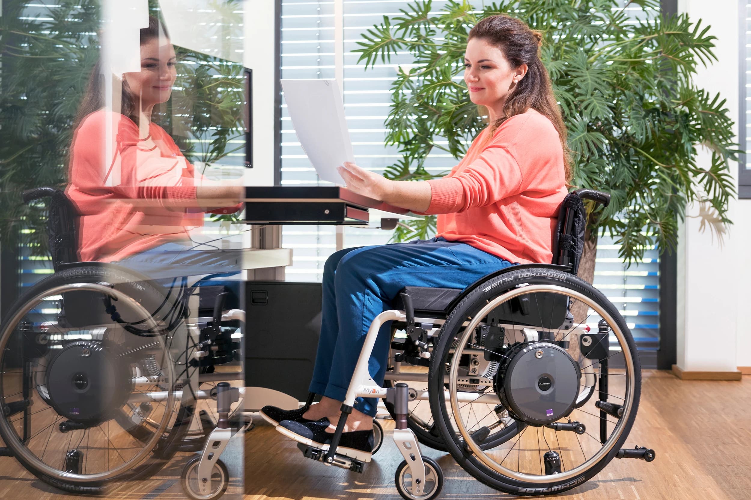 Junge Frau mit langen brünetten Haaren sitzt an ihrem Schreibtisch in ihrem manuellen Rollstuhl mit e-motion Elektroantrieb und liest ein Dokument
