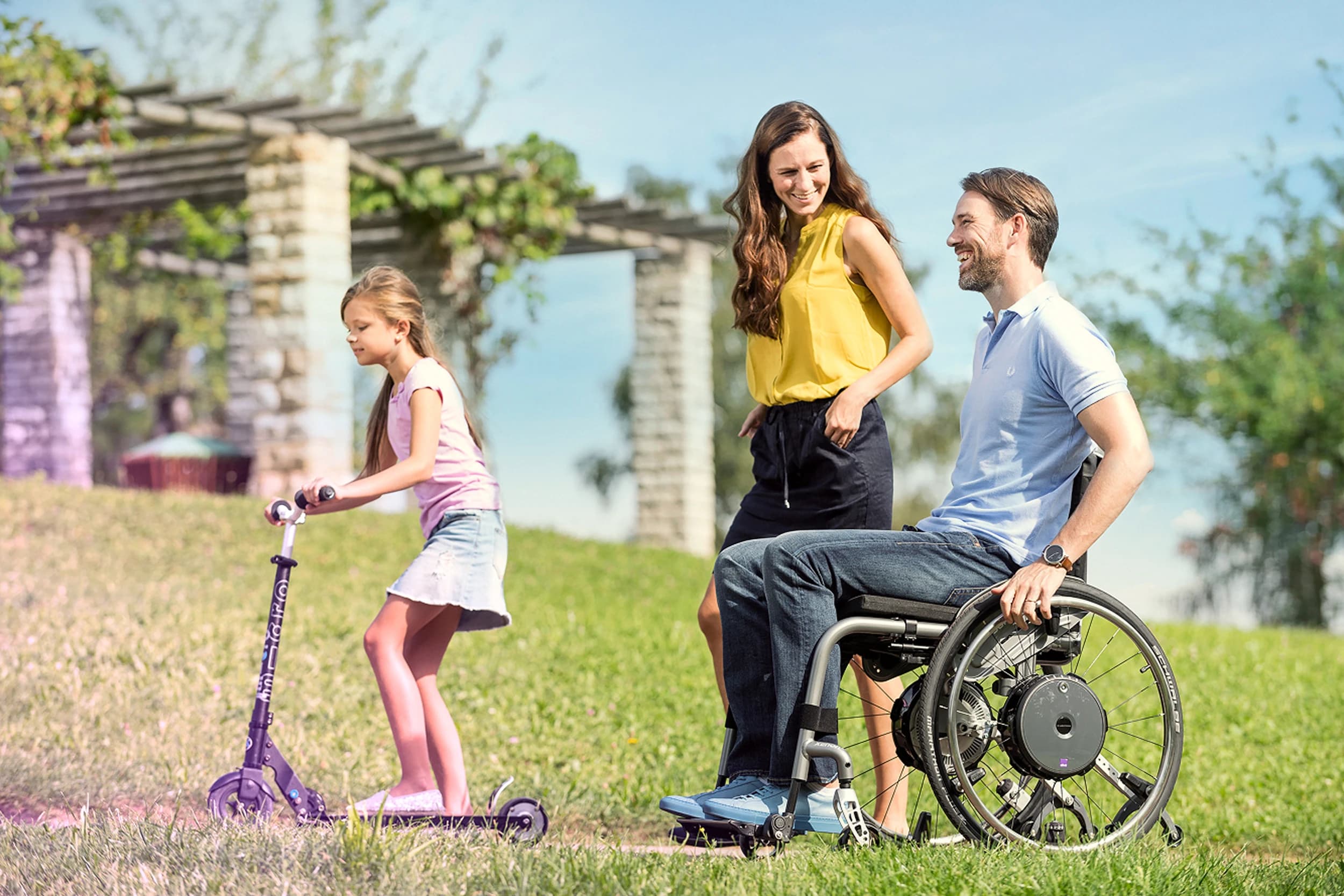 Junge Familie bei der Spazierfahrt mit Papa im manuellen Elektrorollstuhl mit Elektroantrieb e-motion im grünen Park.