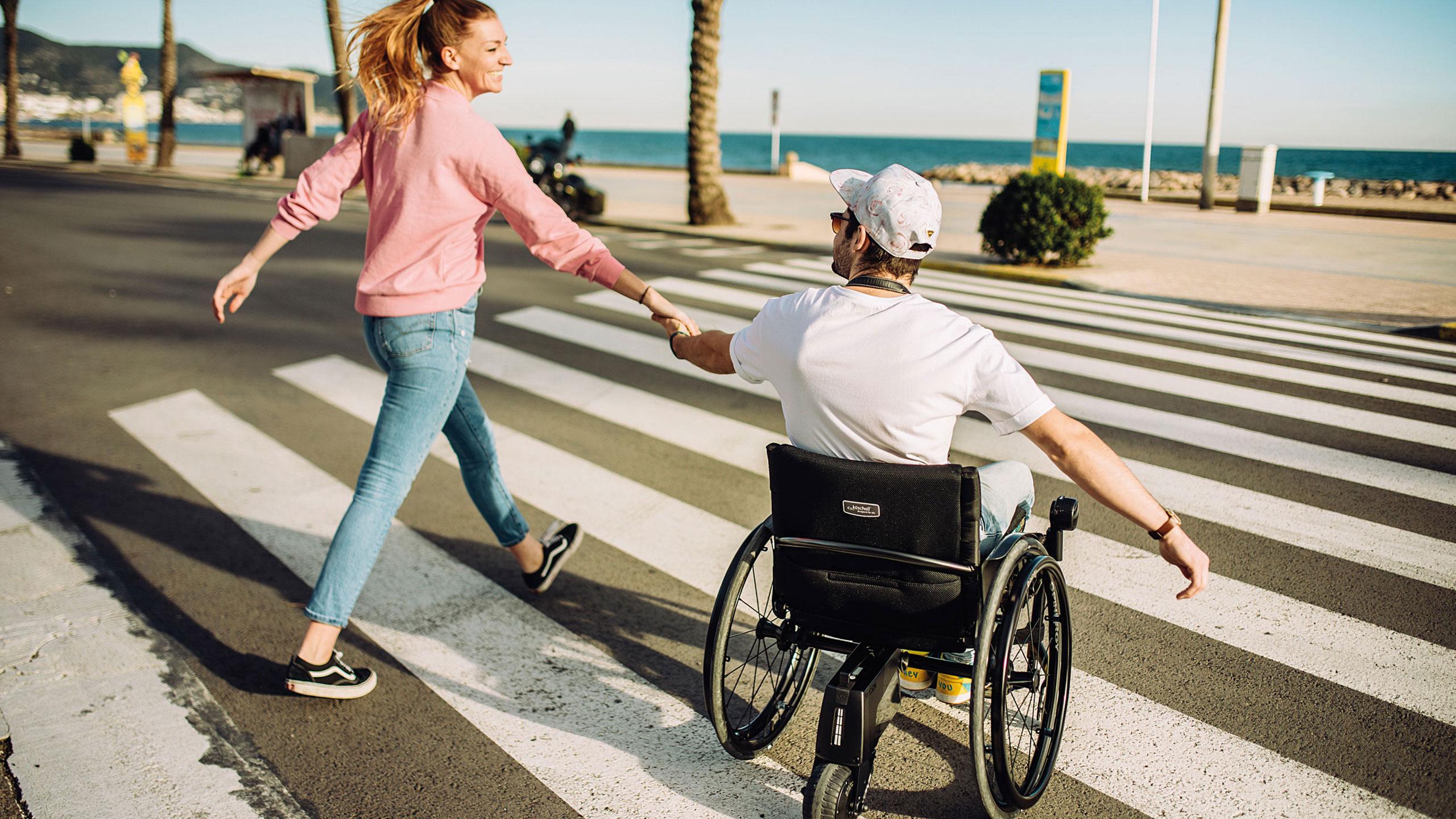 Junger Mann in Aktivrollstuhl mit mit Freundin an der Hand und smoov Elektroantrieb überquert Zebrastreifen vor Barcelona Strandpromenade.