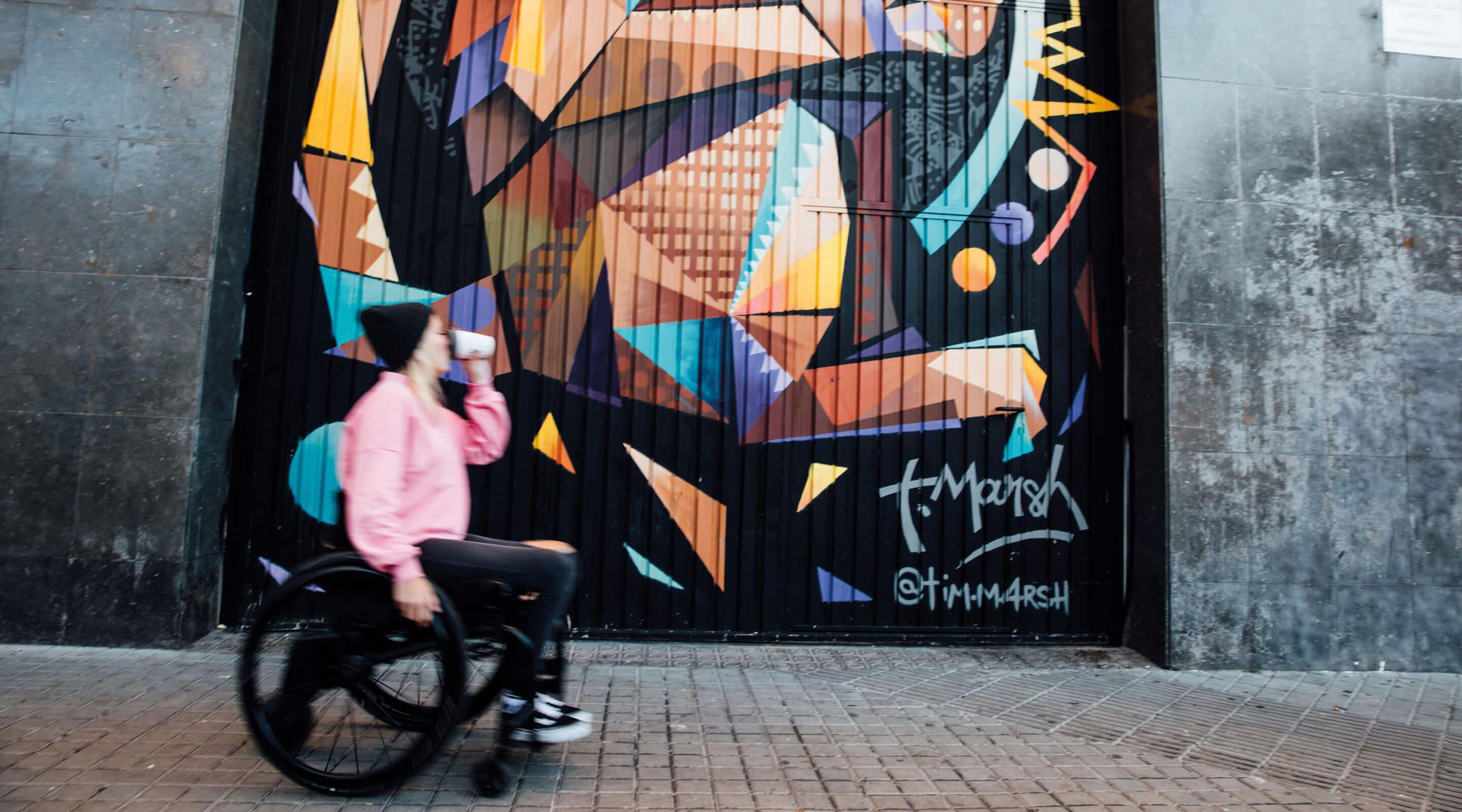 Profilansicht junge Frau in Aktivrollstuhl mit smoov Zusatzantrieb vor Graffiti-Wall und Coffee-to-go Becher in der Hand.