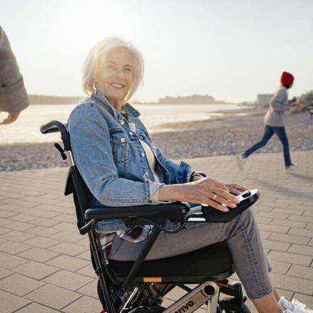 Frau ist am Strand und sitzt lächelnd in Rollstuhl mit Joystick