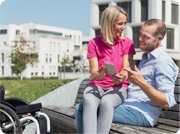 Mann sitzt mit Freundin auf dem Schoß auf einer Bank – vor den beiden ein Rollstuhl mit e-motion Zusatzantrieb – im Hintergrund moderne Gebäude
