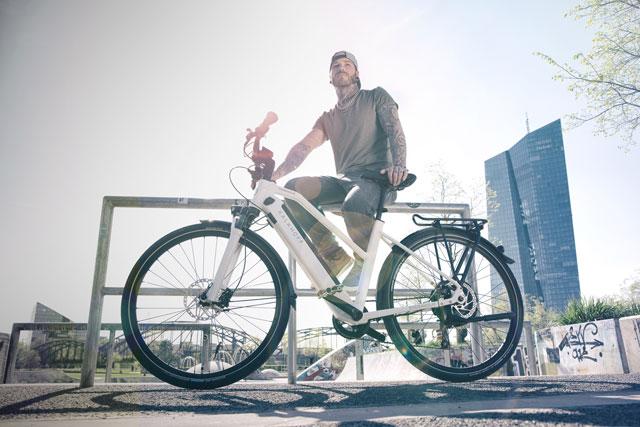 Ein junger Mann mit Basecap sitzt in urbaner Kulisse auf einem Geländer und hält ein Fahrrad mit neodrives E-Antrieb von Alber