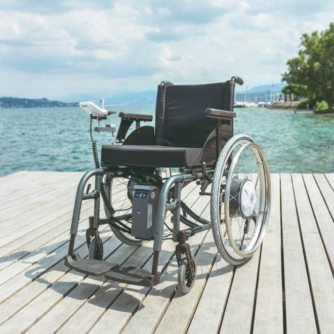 Rollstuhl mit Blick auf alle Komponenten des Zusatzantriebs e-fix steht auf einem Steg am See