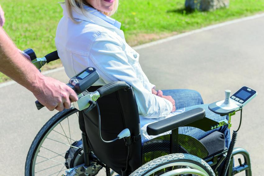 Eine Frau sitzt im Rollstuhl und dreht sich lächelnd zur Kamera. Ein Mann schiebt sie vorwärts.