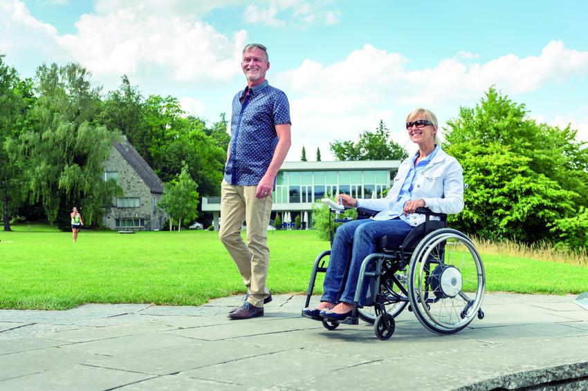 Eine Frau mit Sonnenbrille im Rollstuhl fährt mit ihrem Mann vor einer grünen Wiese spazieren.