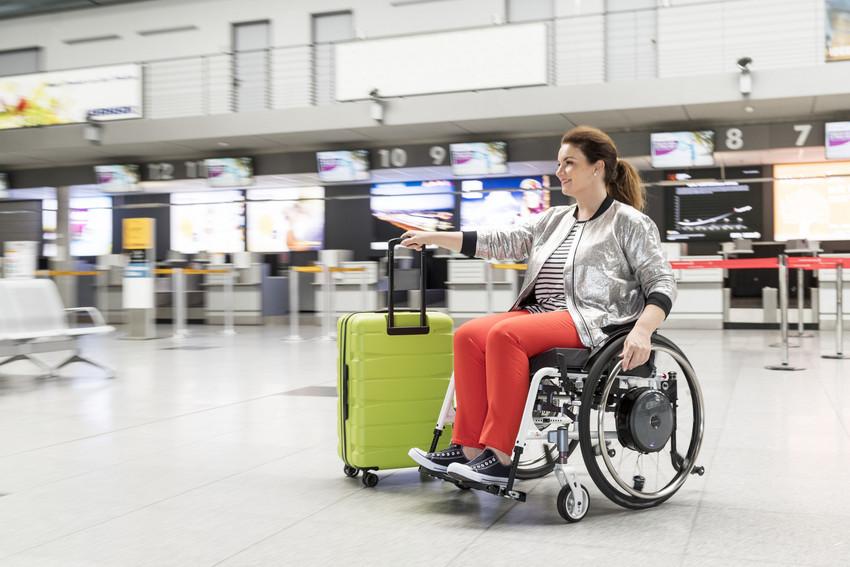 Frau ist am Flughafen mit Rollstuhl und Gepäck in der rechten Hand