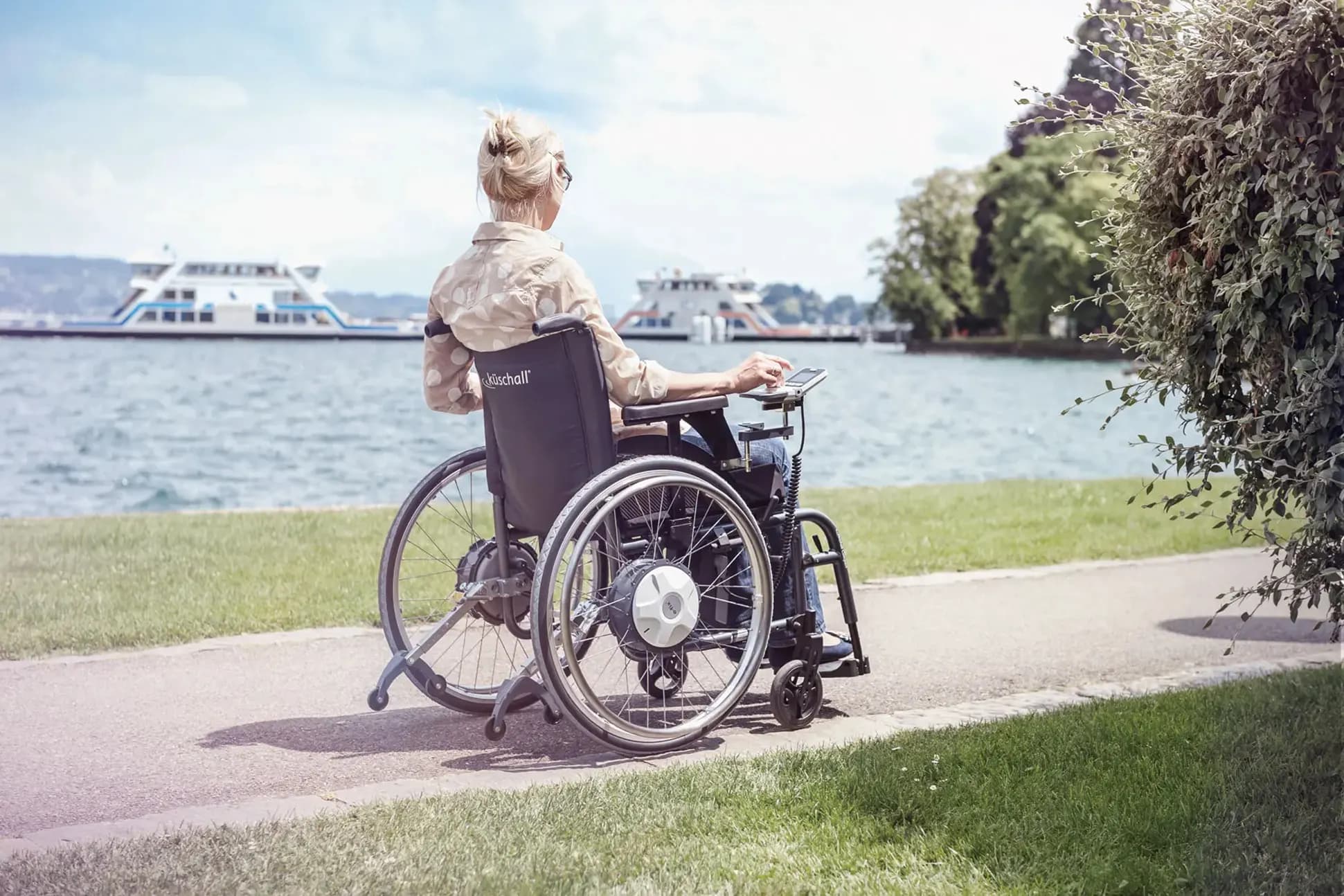 Frau auf Spazierfahrt in ihrem manuellen Rollstuhl mit e-fix Elektroantrieb an der Promenade des Bodensees mit Schiffen im Hintergrund